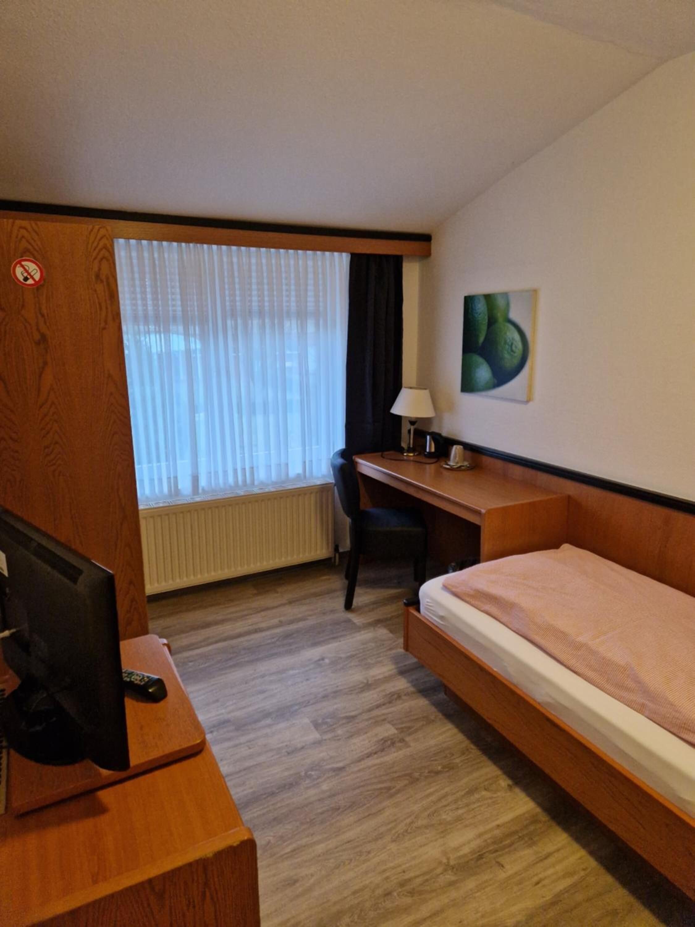 Ein Hotelzimmer mit einem Holztisch, einem Einzelbett, einem Stuhl, einem Fernseher und einem Fenster mit Vorhängen.