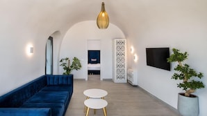A modern living room with a blue sofa, two white tables, a TV, and a potted plant. - Palm Island Suites Santorini (Santorini)