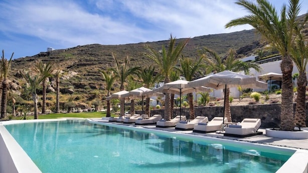 A swimming pool with lounge chairs and umbrellas, surrounded by palm trees and a mountainous backdrop. - Palm Island Suites Santorini (Santorini)