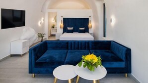 A modern hotel room with a large bed, a blue velvet sofa, two white round tables, and a TV mounted on the wall. - Palm Island Suites Santorini (Santorini)