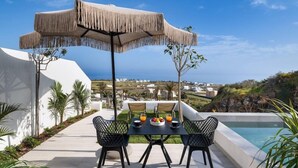 A patio with a table and chairs, a pool, and a view of the landscape. - Palm Island Suites Santorini (Santorini)