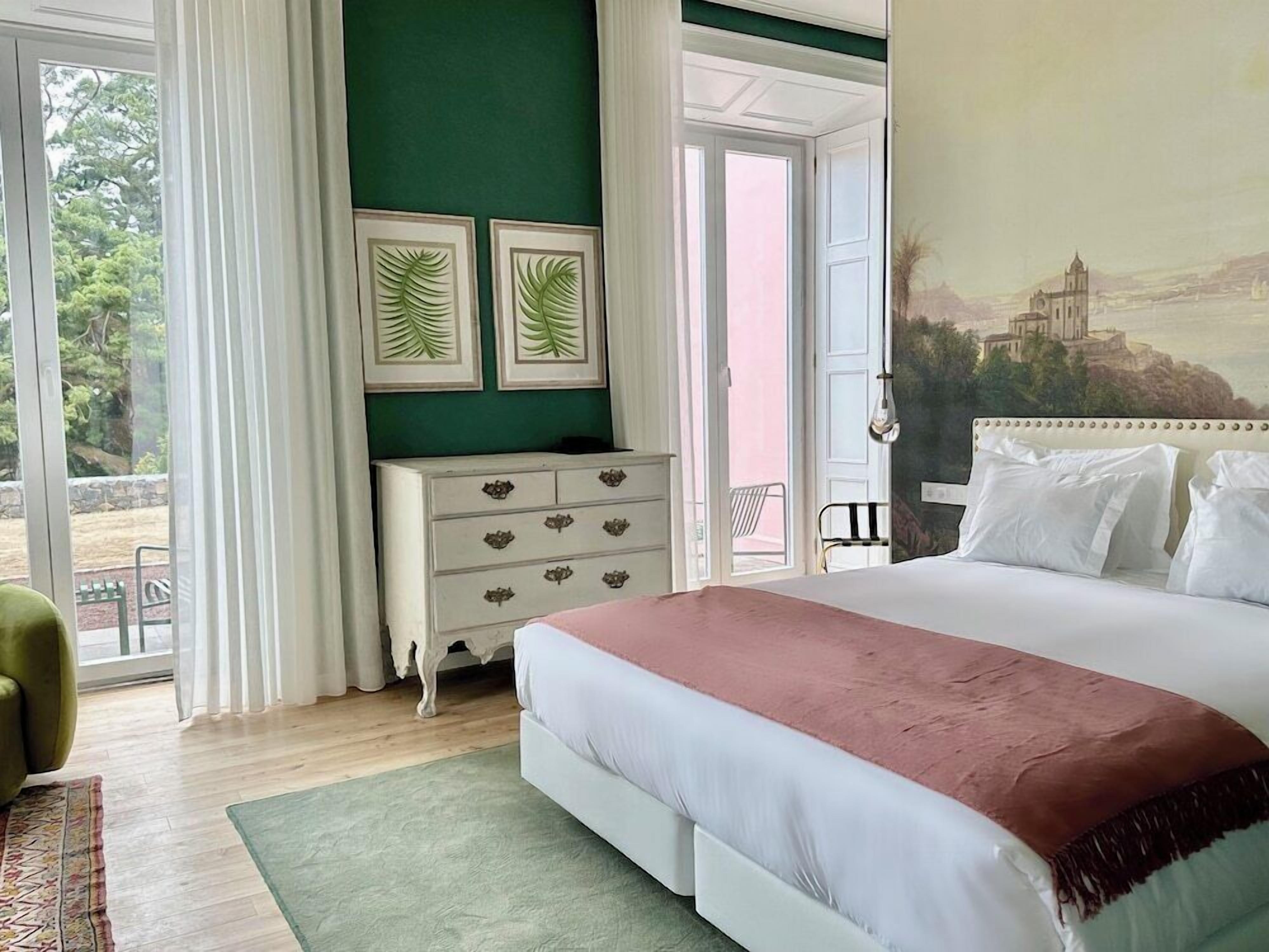 A bedroom with a bed, a dresser, a green wall, and a painting of a building.
