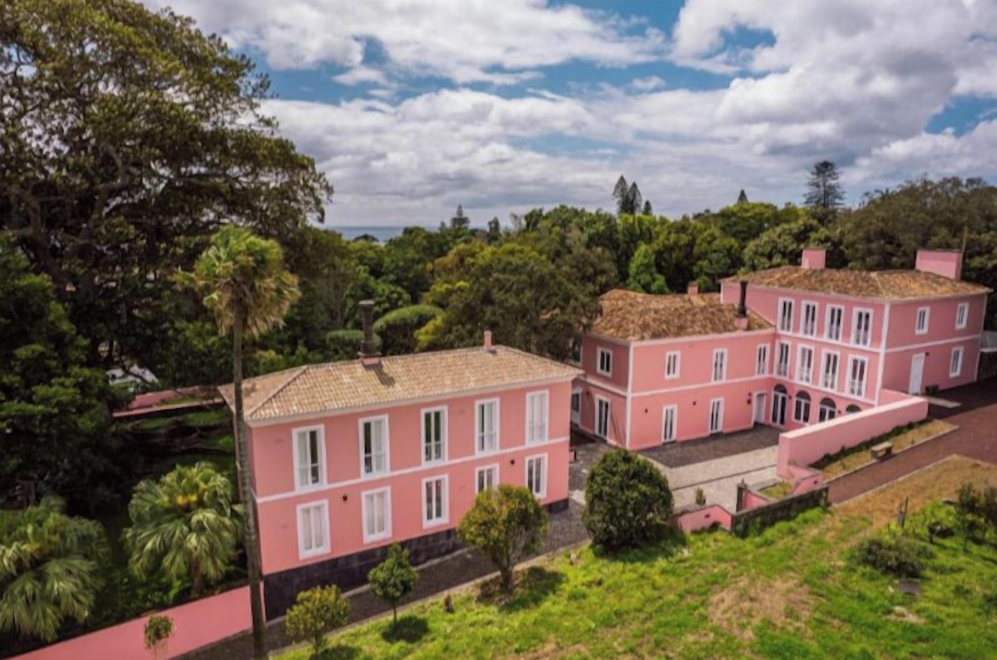 A pink building with multiple windows and a tiled roof, surrounded by greenery.