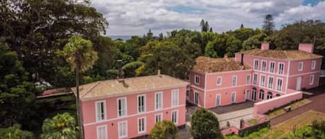 A pink building with multiple windows and a tiled roof, surrounded by greenery.