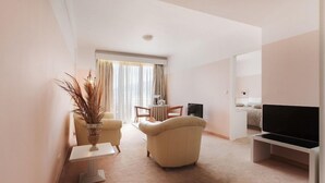 A modern living room with a sofa, a coffee table, and a TV stand. There is a dining area with a table and chairs. The room has a large window with curtains. - Hotel Z Palace and Congress Center (Xanthi)