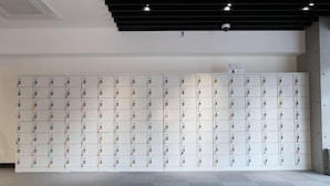 A row of white lockers with colored tags, a security camera, and a sign on the wall. - Lightning Hotel (Tokyo)