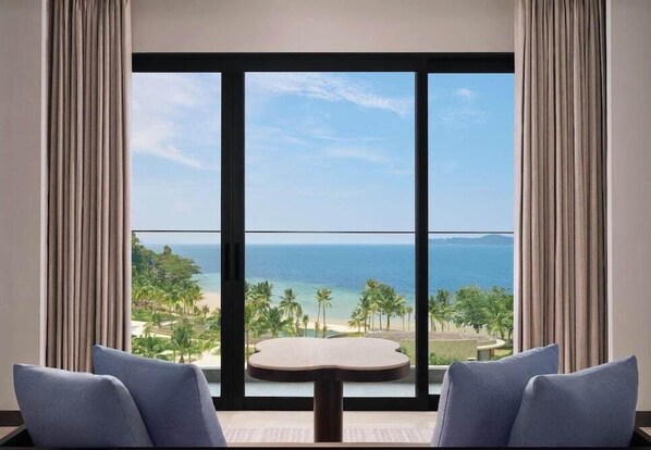 A room with a large window offering a view of a beach and ocean. There is a round table and two chairs in the room. - Lampung Marriott Resort and Spa (Bandar Lampung)