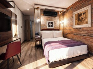 Un dormitorio moderno con pared de ladrillo, cama con manta morada, escritorio con silla y televisor montado.