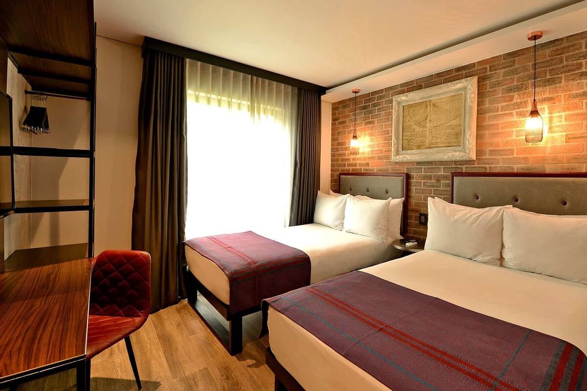 A hotel room with two beds, a desk, a chair, and a brick wall with a framed picture.