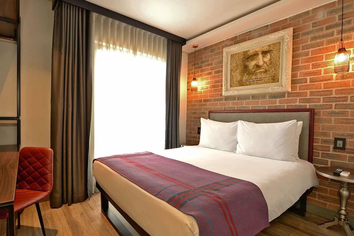 A hotel room with a bed, a desk, a chair, a window with curtains, a brick wall, and a framed picture.