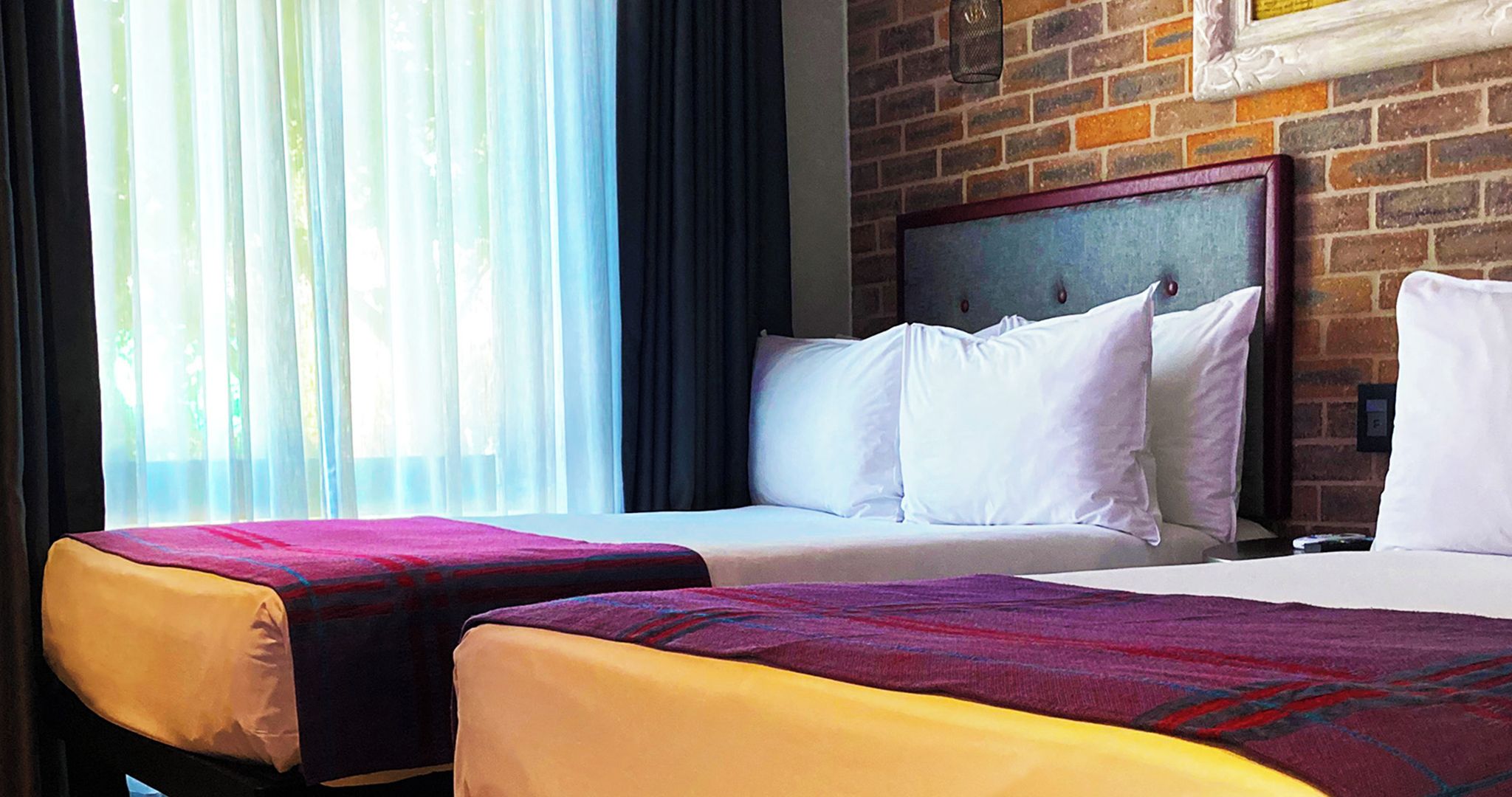 Quarto de hotel com duas camas, uma parede de tijolos e uma janela com cortinas.