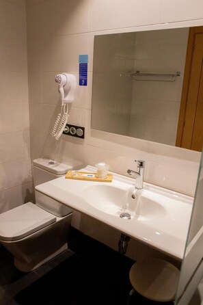 Une salle de bain avec un lavabo blanc, un miroir, un sèche-cheveux, des toilettes et un petit tabouret.