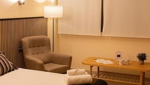 Een hotelkamer met een groot bed, een stoel, een tafeltje met een lamp en een raam met jaloezieën.