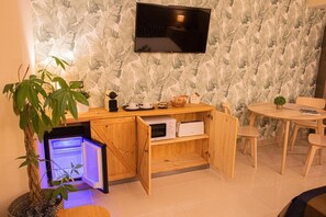 Una habitación compacta con un mueble de madera, un refrigerador pequeño, un televisor montado en la pared y una zona de comedor con mesa y sillas.