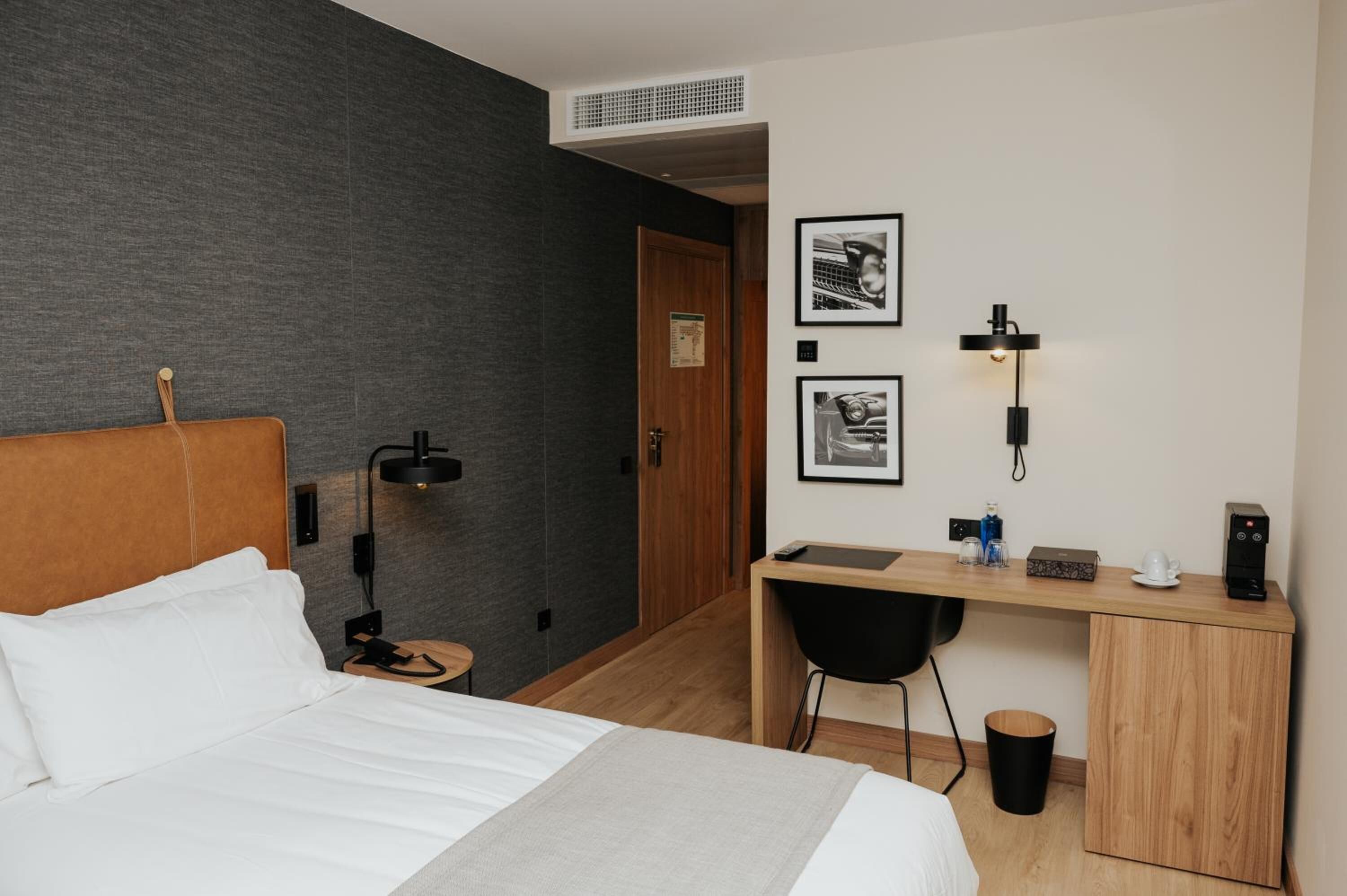 A modern hotel room with a bed, desk, and chair, a small table with a lamp, and a wall-mounted shelf with a picture frame and a lamp.