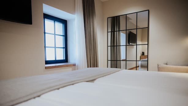 Quarto de hotel com cama, janela com cortinas, espelho e uma cadeira.
