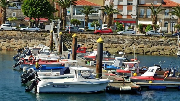 Un port de plaisance où sont amarrés différents types de bateaux, un mur de pierre et, au fond, des bâtiments aux toits rouges.
