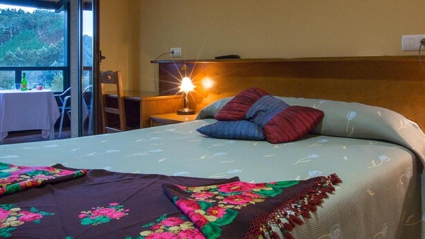 A bedroom with a bed, a nightstand, a lamp, and a view of the outdoors. - Hotel Restaurante Canero (Valdes)