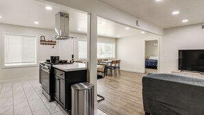 Et moderne køkken med komfur, vask og skraldespand i rustfrit stål. Der er et spiseområde med et bord og stole, og en stue med sofa og fladskærms-TV.