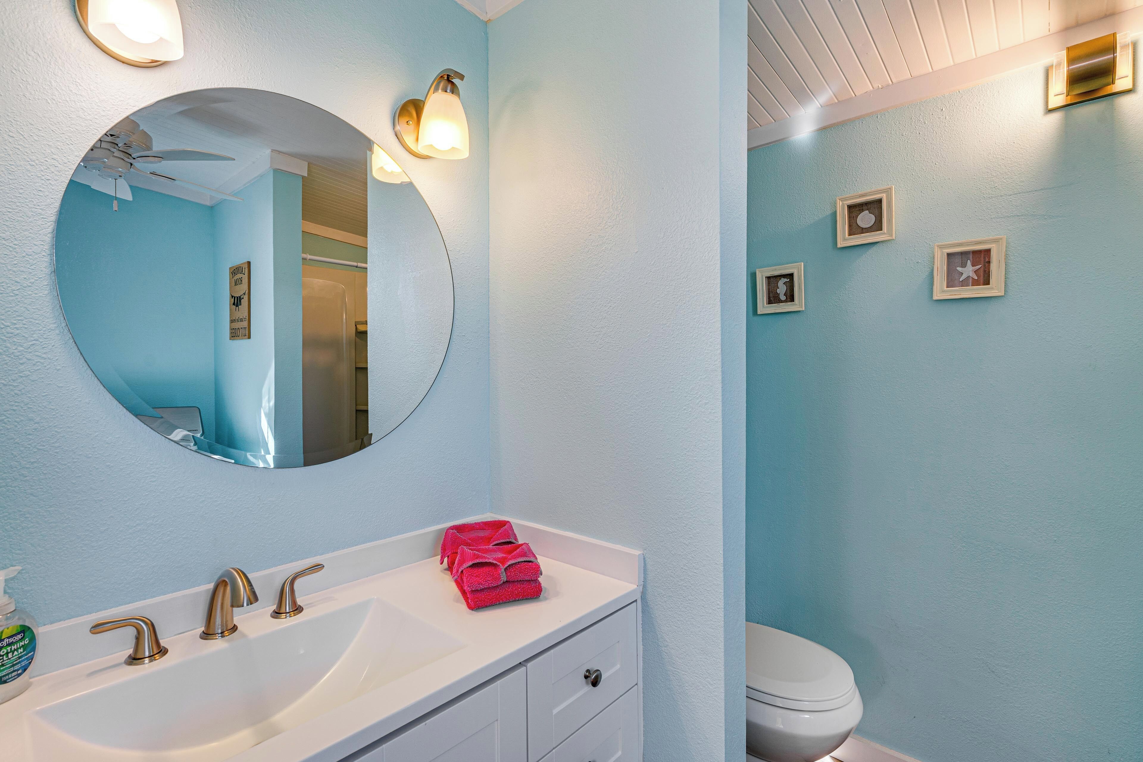 A bathroom with a white sink, a round mirror, and a toilet. There are framed pictures on the wall and a ceiling fan.