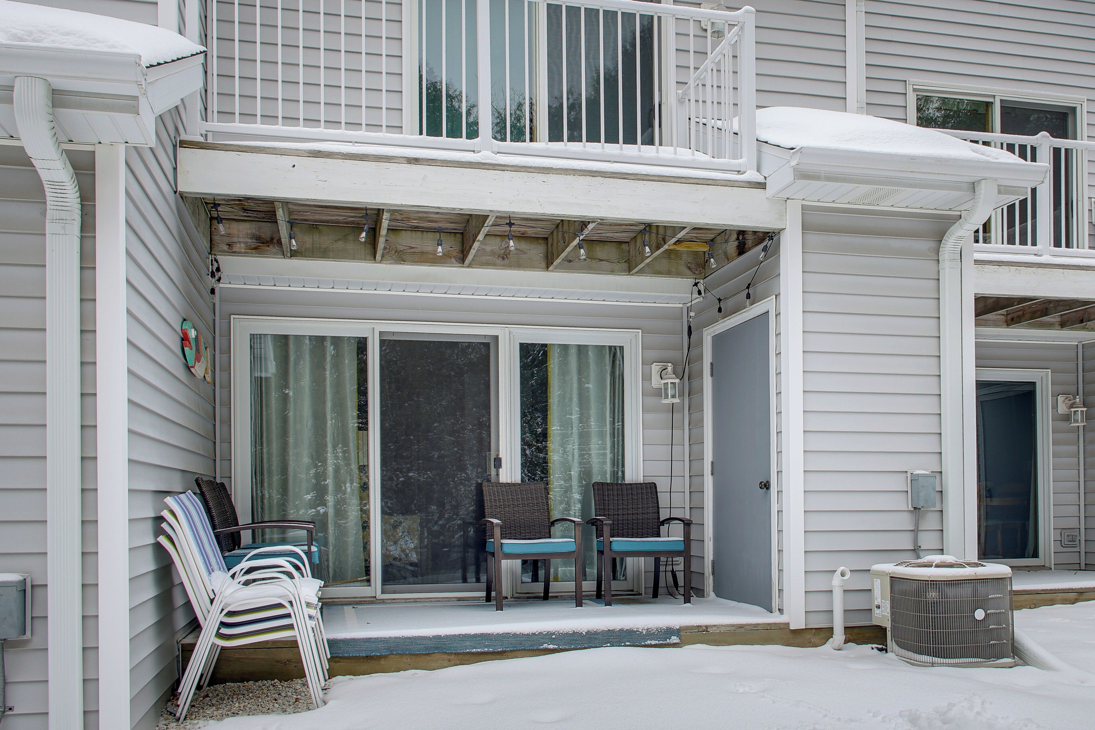 A covered porch with two chairs and a small table, a sliding glass door, and an air conditioning unit on the ground.