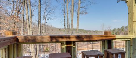 Eine Holzdielen-Terrasse mit drei Hocker und einem Tisch, mit Blick auf eine verschneite Landschaft und Bäume.