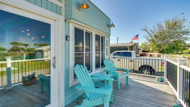 Um deck de madeira com duas cadeiras Adirondack e uma mesa, uma área coberta com vista para a praia, e um veículo estacionado por perto.