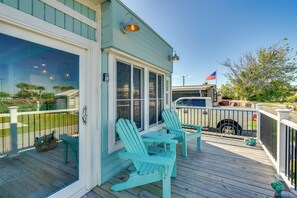 Una terraza de madera con dos sillas Adirondack y una mesa, un área cubierta con vista a la playa, y un vehículo estacionado cerca.
