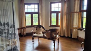 Een kamer met een bed, overdekt door een dunne gordijn, twee houten stoelen, een kleine tafel en grote ramen met gordijnen.