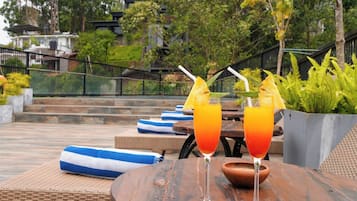Dos vasos de bebida de naranja sobre una mesa de madera, con vistas a un jardín y edificios al fondo.