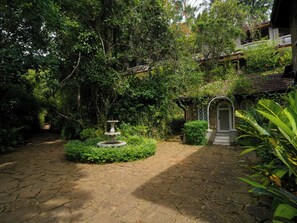 A garden with a stone pathway leading to a central fountain, surrounded by lush greenery and a stone building with a balcony. - Graystone Bungalow by Seven Angels (Diyatalawa)