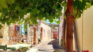 A narrow street with stone buildings, outdoor seating, and a tree providing shade. - Family 5BR Villa with sea views, private pool, summer kitchen & spacious garden (Almyrida)