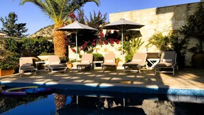 A poolside area with lounge chairs and umbrellas, surrounded by plants and a stone wall. - Family 5BR Villa with sea views, private pool, summer kitchen & spacious garden (Almyrida)