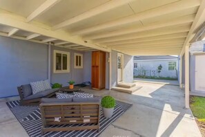 Eine überdachte Terrasse mit einer Couch, einer Holzbank und einem kleinen Tisch. Es gibt Fenster und eine Tür zum Haus.