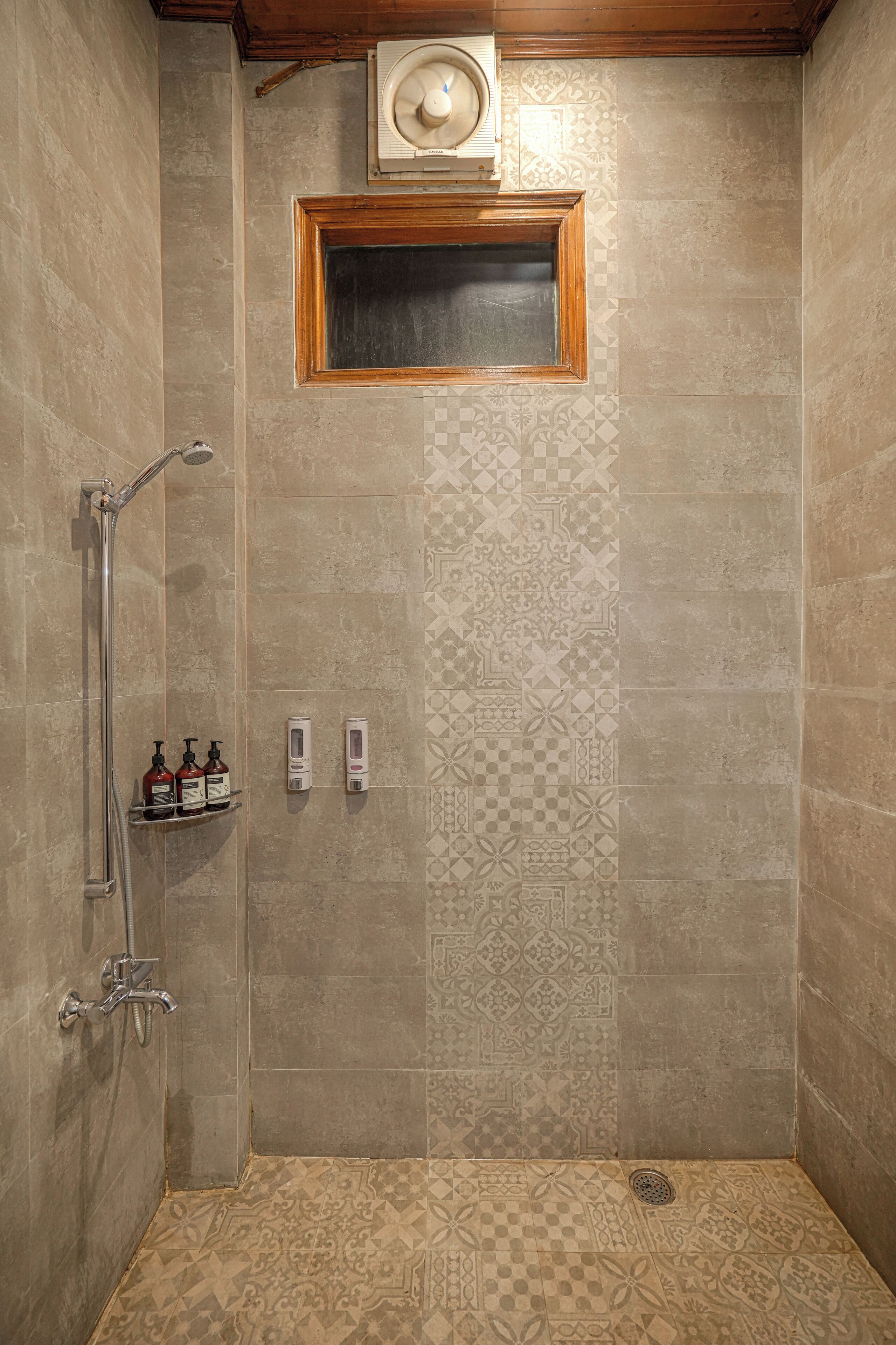 Ducha a pie con azulejos estampados, cabezal de ducha manual, ventilador de pared y dos dispensadores de jabón.