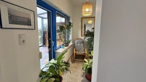 Un pasillo estrecho con una puerta azul que da a un espacio exterior, una silla y varias plantas en macetas.