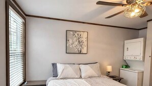Ein Schlafzimmer mit einem Bett, einem Deckenventilator, einem Fenster mit Jalousien, einem Wandbild und einem kleinen Tisch mit einer Lampe.