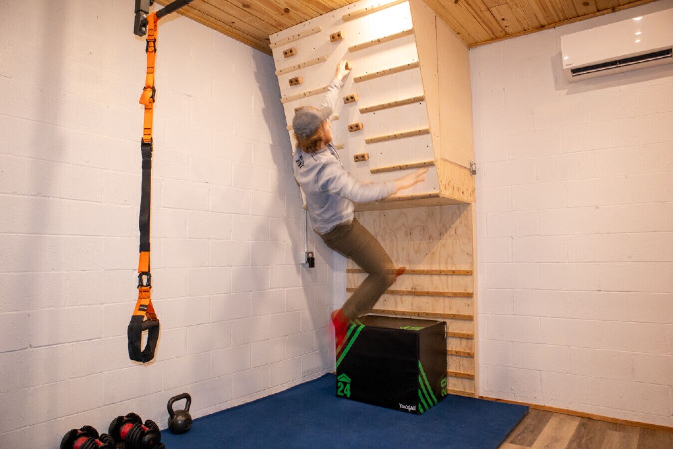 Iemand beklimt een houten muur met handgrepen, een zwarte doos met groene strepen en een witte airconditioner aan de muur.