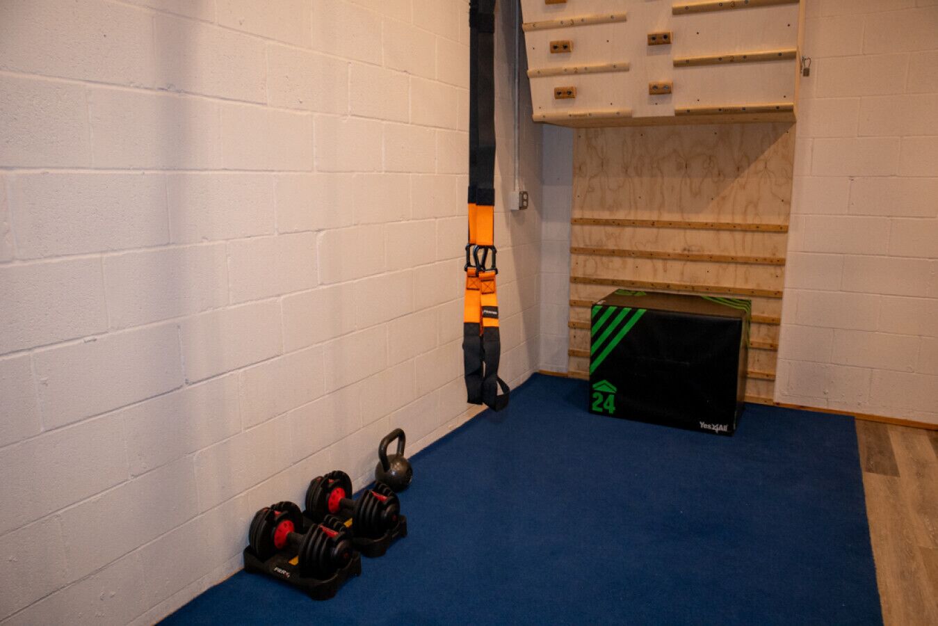 A room with white brick walls, a blue carpet, a kettlebell, a jump rope, and a black box with green stripes.