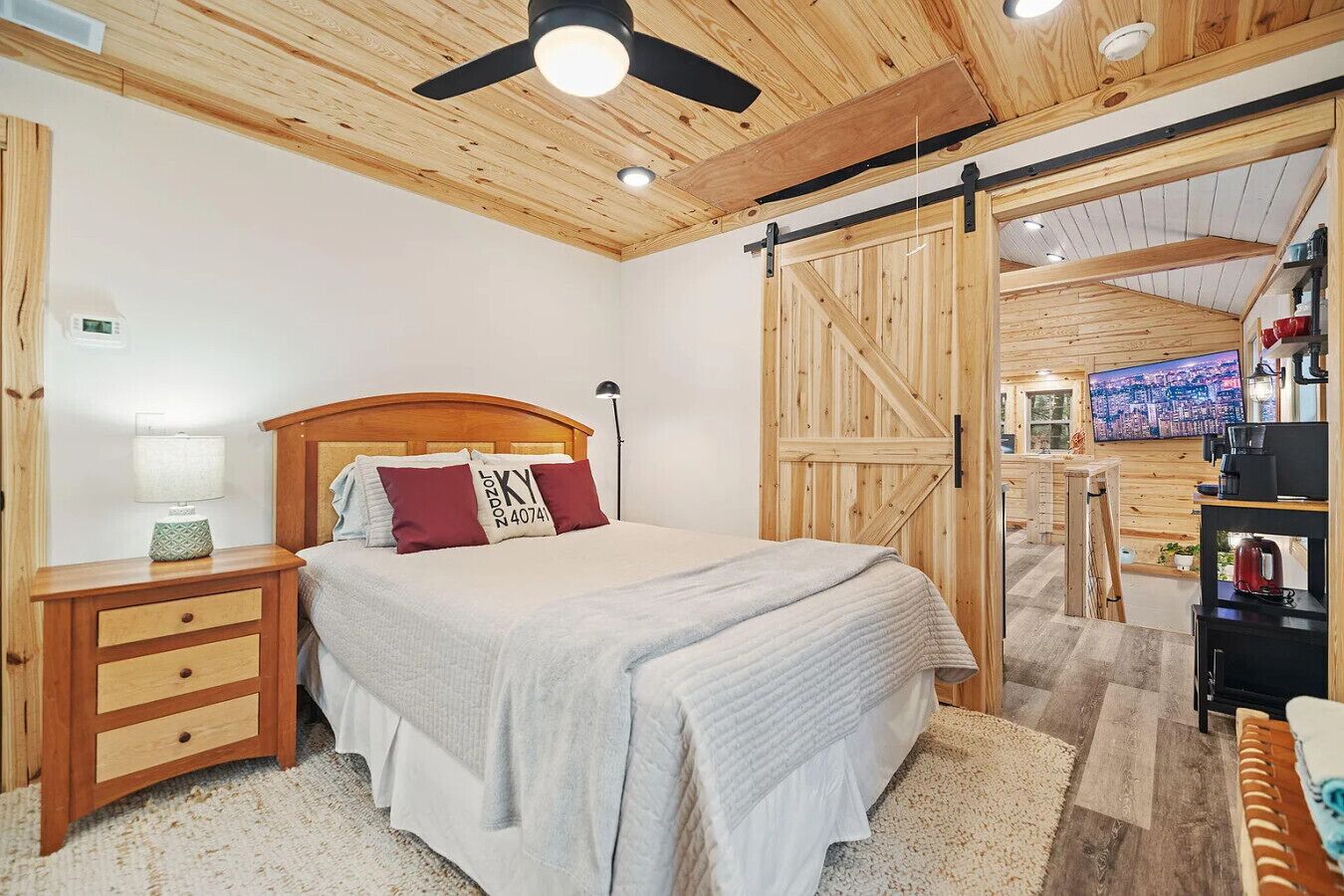 A bedroom with a wooden ceiling, a bed with a headboard, a nightstand, a lamp, and a TV in the background.