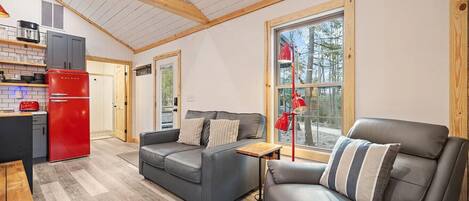 A compact living space with a kitchenette, a gray sofa, a leather armchair, and a large window offering a view of the outdoors.