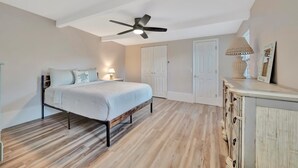 Ein Schlafzimmer mit einem Bett, einem Deckenventilator, einem Nachttisch, einer Lampe, einer Kommode und einer Tür.
