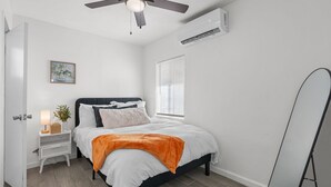 A bedroom with a bed, bedside table, nightstand, ceiling fan, air conditioning unit, window, and a standing mirror. - Casa Esmeralda · Cozy · Midtown Gem (Tucson)