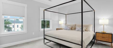 A bedroom with a four-poster bed, a nightstand with a lamp, and a window with blinds.