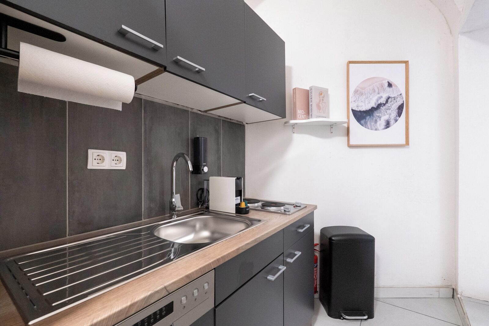 Een moderne keuken met een roestvrijstalen spoelbak, een inbouwoven en grijze keukenkastjes. Er hangt een zwarte speaker en een lijst met een afbeelding aan de muur.