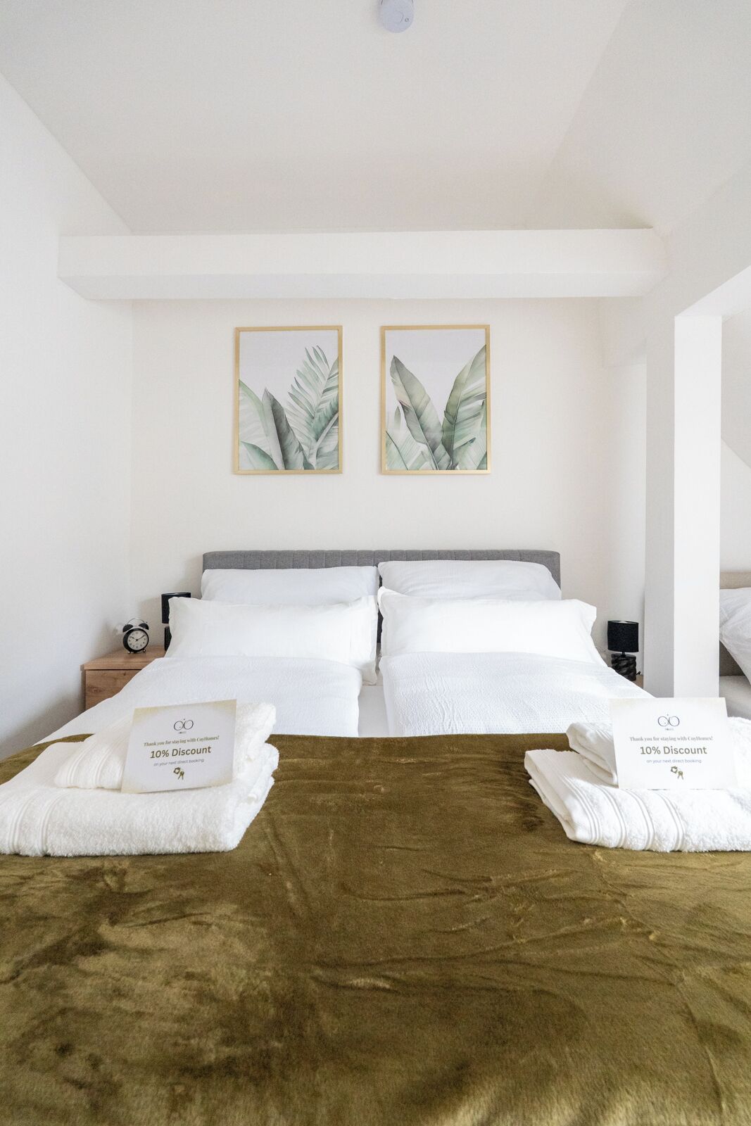 A hotel room with a double bed, two framed botanical prints on the wall, a bedside table with a lamp, and a sign offering a 10% discount on direct bookings.