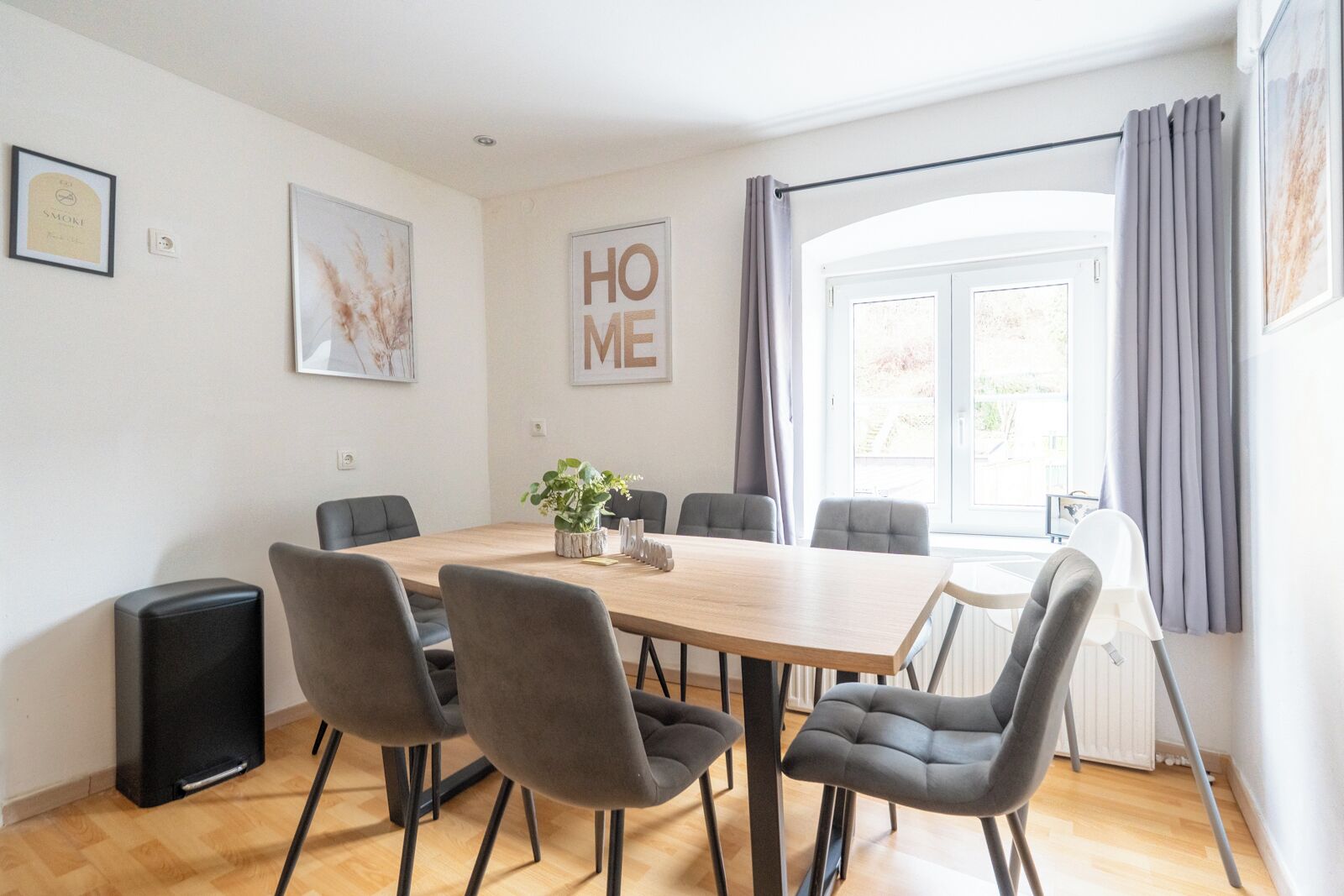 Un comedor moderno con una mesa de madera, sillas tapizadas en gris y un ventanal con cortinas grises.