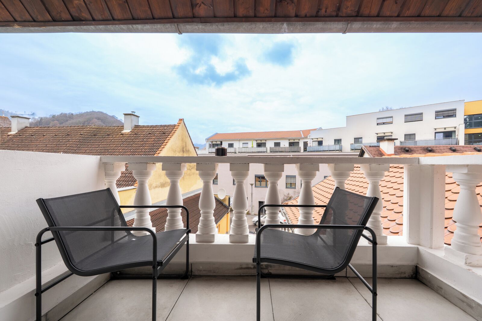 A balcony with two black chairs, white balustrades, and a view of buildings with red-tiled roofs.
