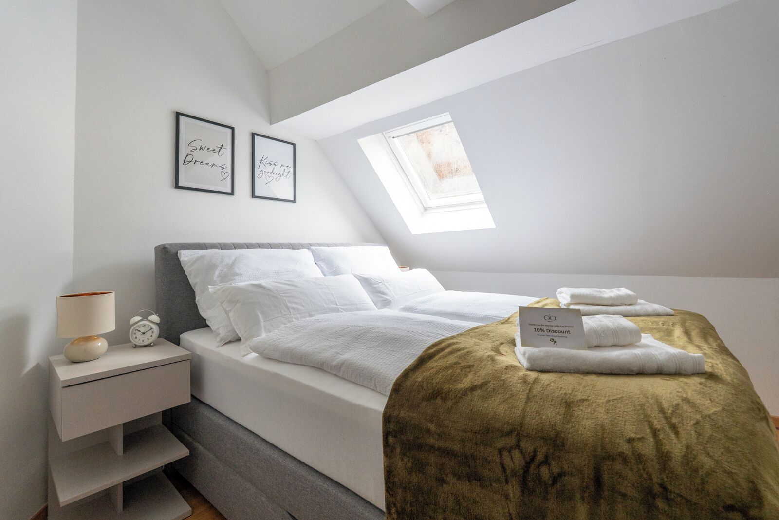 Un dormitorio ordenado con cama, mesita de noche, lámpara y citas inspiradoras enmarcadas en la pared.