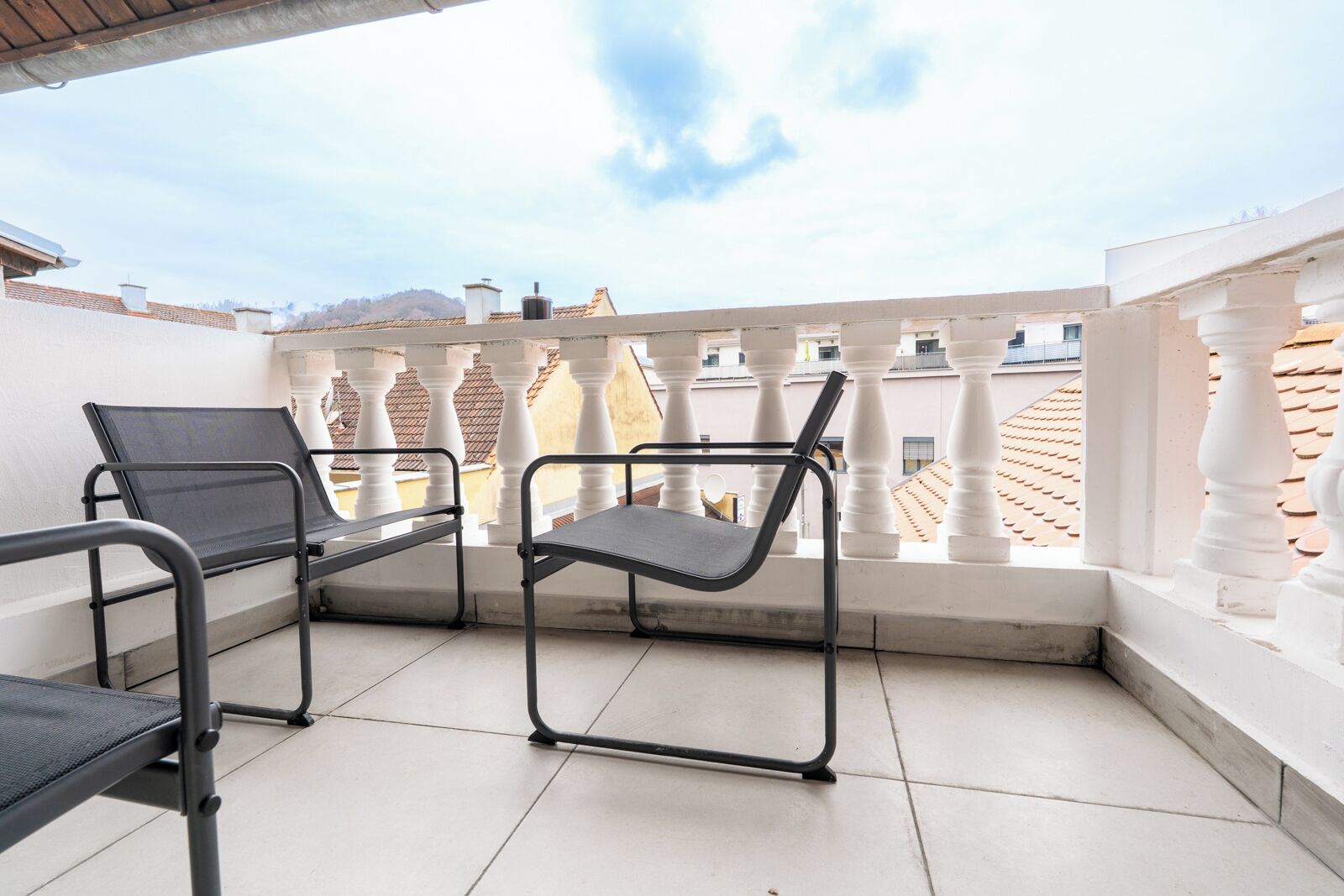 A balcony with two black chairs and a table, white balustrades, and a view of neighboring buildings.